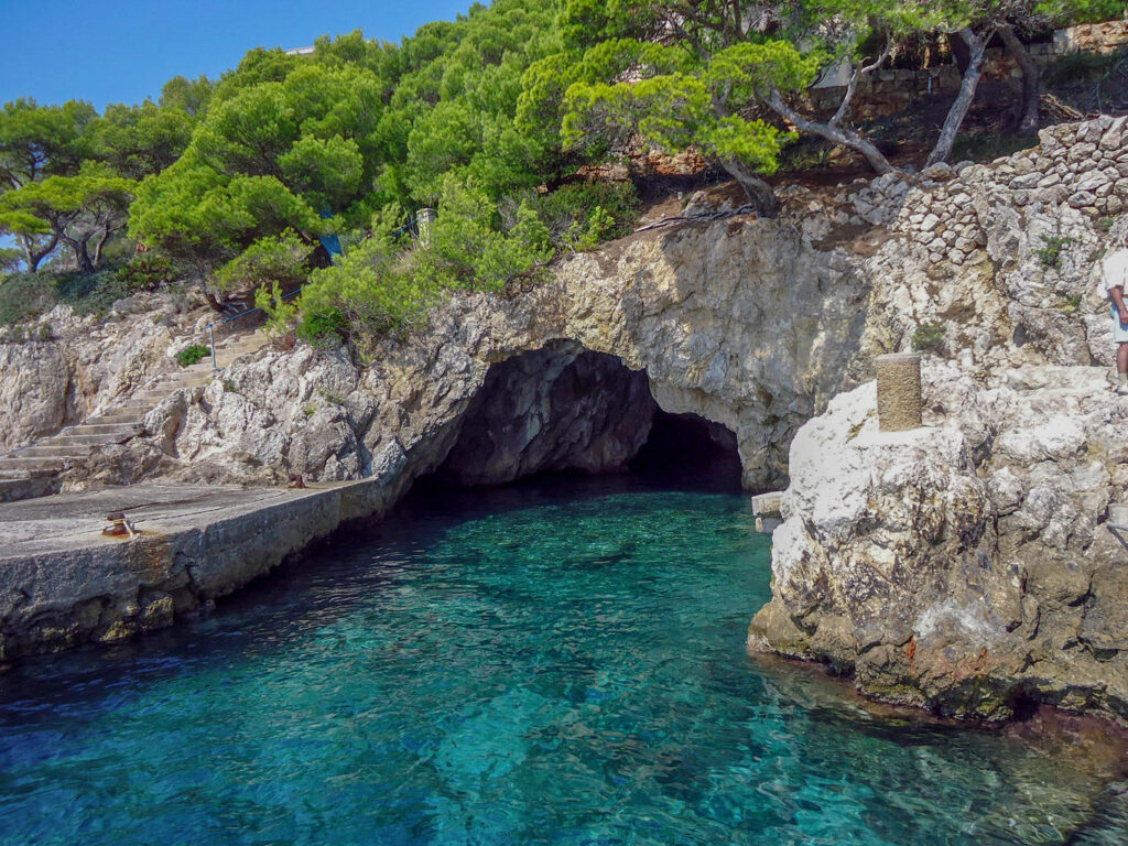 Küstenfoto mit leuchtendgrüner Pinie und Treppe zum Anleger in türkisblaue Bucht mit Steinhöhle im Osten auf Mallorca