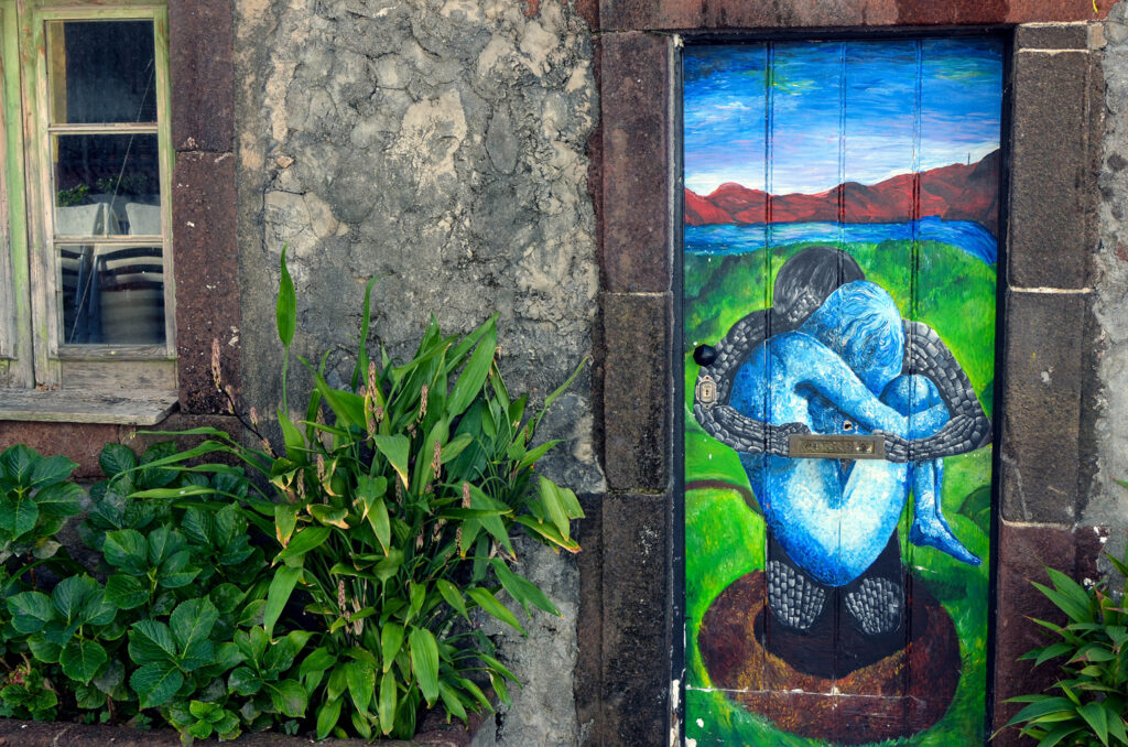 Kunstwerk auf Haustüre mit zwei umschlungenen Wesen in blau und grau auf einer Wiese mit Meer und Gebirge im Hintergrund