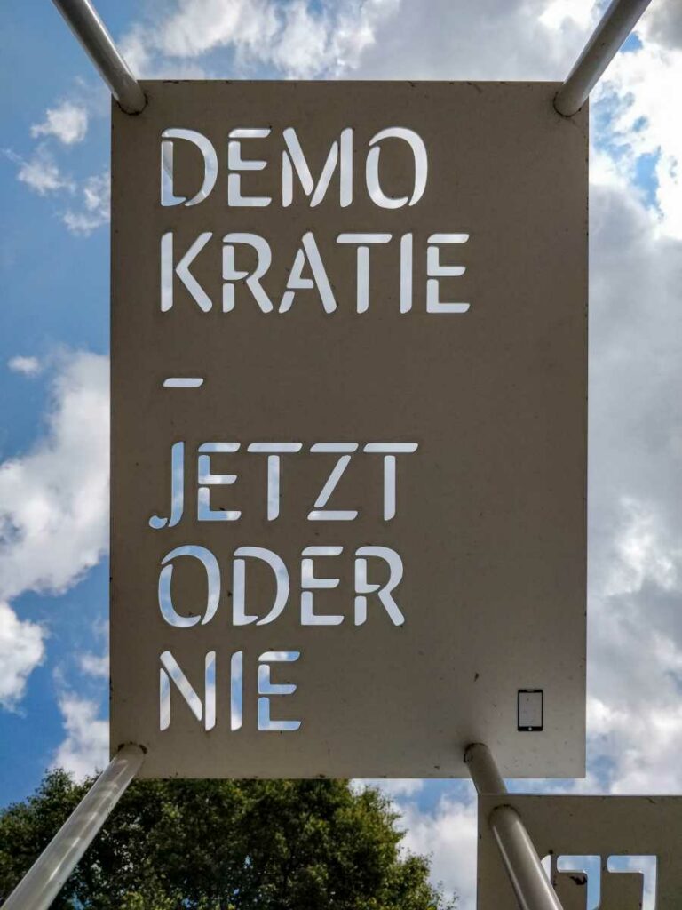 Kunst an der Kirche in Waren Müritz. Metalltafel wo man den Spruch Demokratie - Jetzt oder nie lesen kann, wenn man in den Himmel sieht