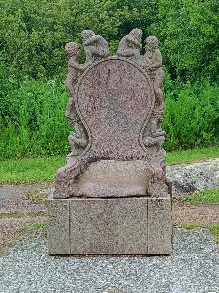 aus Stein gemeißelter Sessel auf einem Block mit Figuren um den Rücken herum, Skulpturenpark Ploumanach