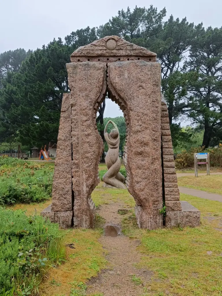 ein Tor aus Granitsteinen, der Durchblick gibt die Frauenfigur mit verschränkten Beinen, hoch zusammengeführten Armen und gesenktem Kopf frei, Skulpturengarten Ploumanach